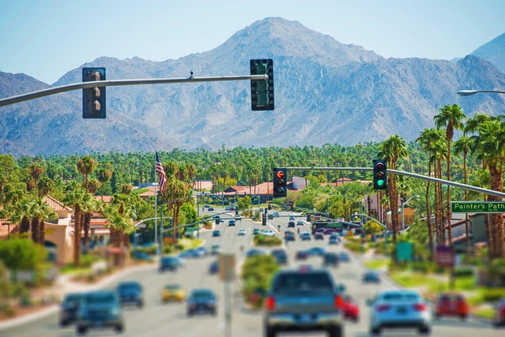 a vibrant, bustling scene of attendees enjoying gourmet food and exquisite wine at the palm springs food and wine festival, set against an elegantly decorated indoor venue with colorful displays and a festive atmosphere. A busy street scene in Palm Springs unfolds with cars stopped at a traffic light. Lush palm trees line the road, and a mountain range rises in the background under the clear blue November sky.