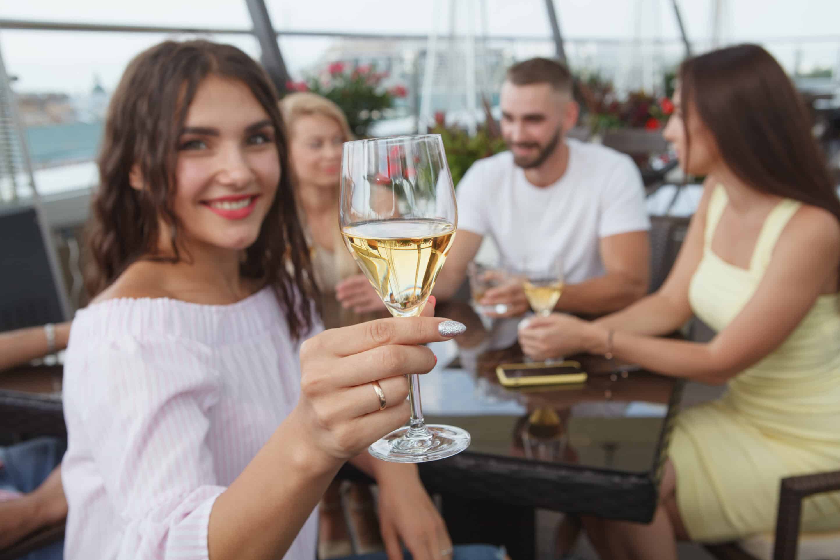 A woman in a light purple top holds up a glass of white wine toward the camera, smiling. Three people sit around the table with her, engaged in conversation, with drinks in hand. The setting appears to be an outdoor patio, perhaps savoring a chef's fall menu on a crisp November evening.