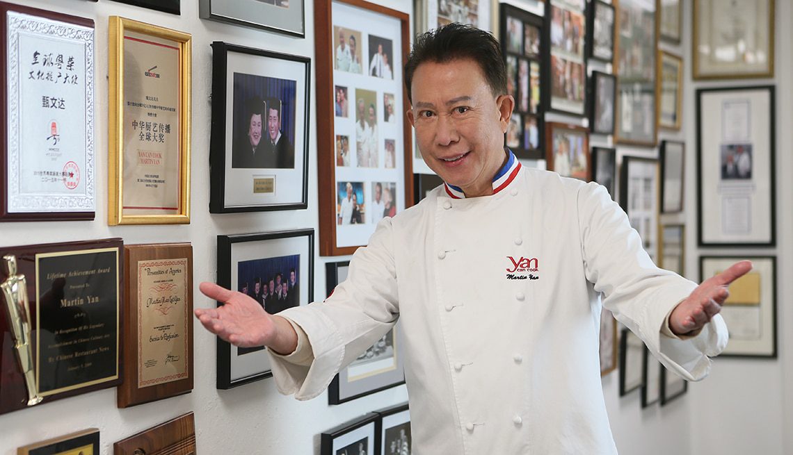 A chef in a crisp white uniform poses with outstretched arms, smiling in front of a wall filled with awards and framed photos, while holding a glass of fine wine.