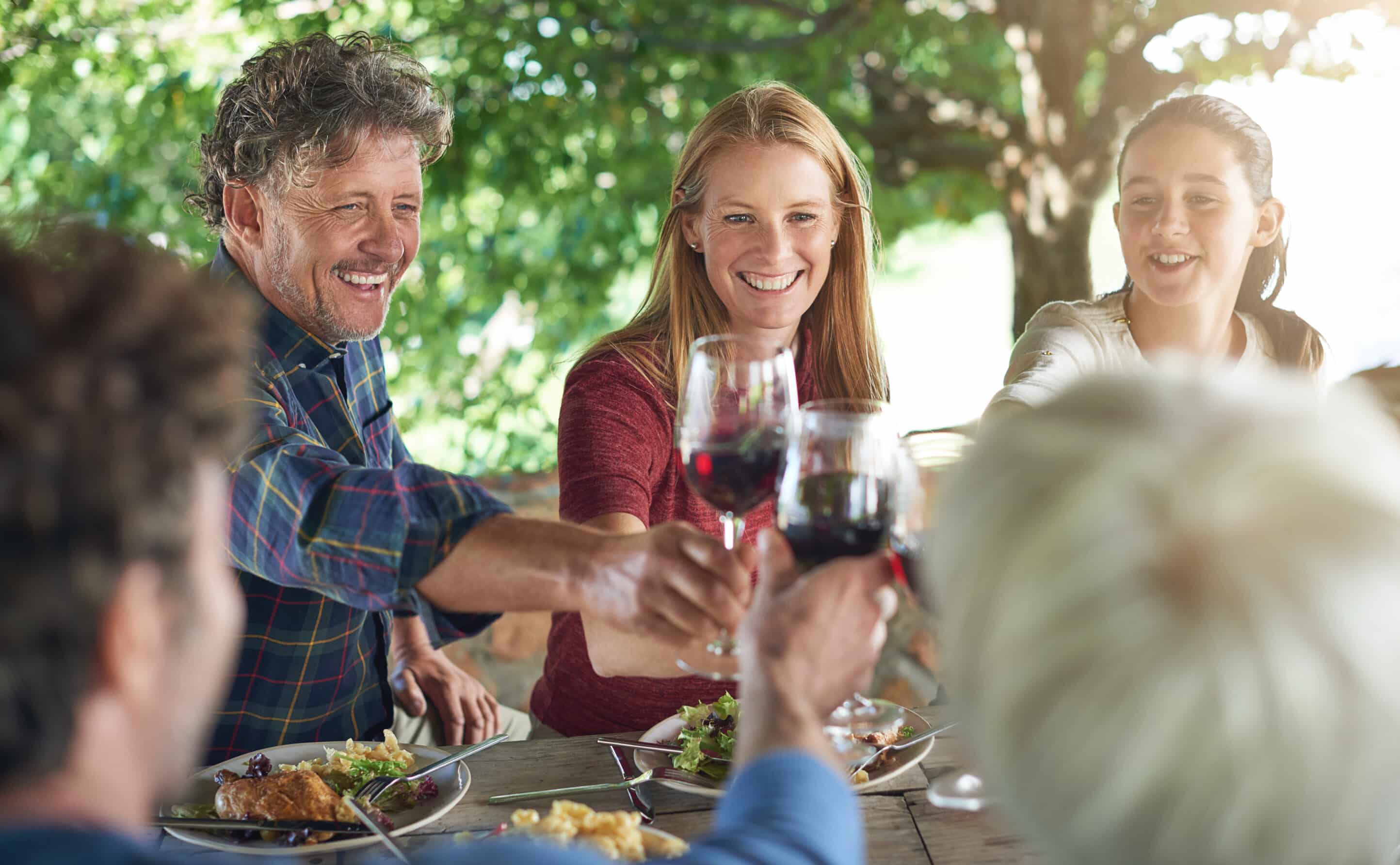 A group of people sitting outdoors at a table under a tree in Palm Springs, toasting with glasses of red wine. They are smiling and sharing a gourmet meal, creating a joyful and lively atmosphere. The background is blurred, highlighting the focus on the group and their togetherness.