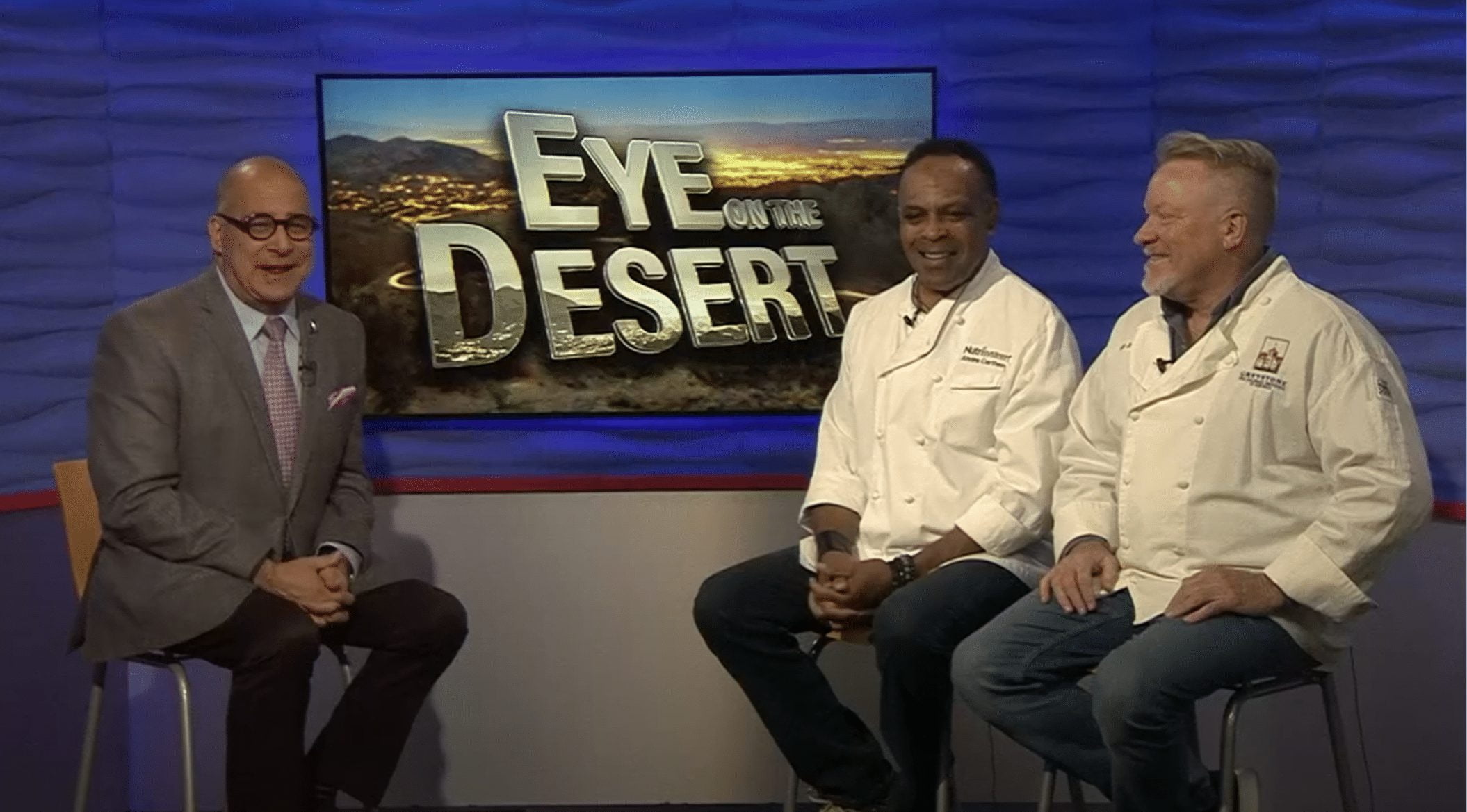 A TV show set with three men sitting in front of a screen displaying "Eye on the Desert." Two are wearing white chef jackets, likely sharing their culinary expertise alongside wine pairings. The third man is in a suit with glasses, all framed by a blue patterned wall.