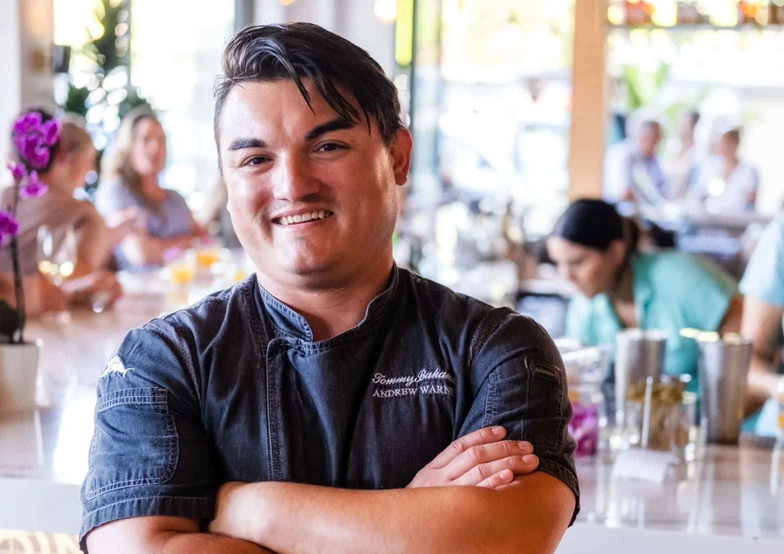 A person wearing a chef's coat smiles while standing with arms crossed in a bright, bustling Palm Springs restaurant. There are people sitting and conversing in the background, enjoying wine. Orchids are visible on the counter, adding to the inviting November ambiance.