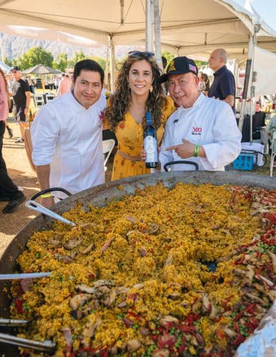 A large paella pan filled with vibrant rice, vegetables, and meat is in the foreground. Behind it, two gourmet chefs in white coats and a woman in a yellow dress smile at an outdoor event. One chef holds a bottle of wine as they stand under a tent in sunny Palm Springs.