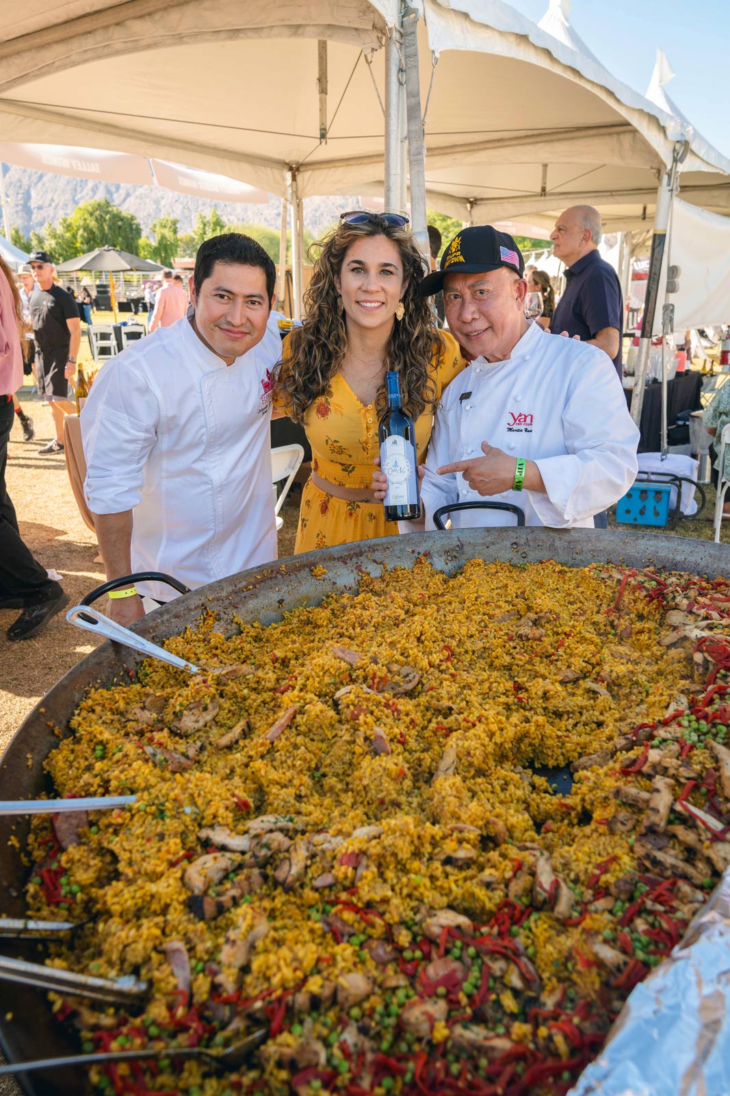 A large paella pan filled with vibrant rice, vegetables, and meat is in the foreground. Behind it, two gourmet chefs in white coats and a woman in a yellow dress smile at an outdoor event. One chef holds a bottle of wine as they stand under a tent in sunny Palm Springs.