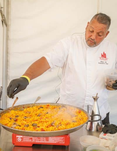 A gourmet chef in a white coat and black gloves is adding ingredients to a large pan of sizzling paella on a portable gas stove. The chef stands in a white tent with various cooking items around, including bowls of chopped ingredients, a metal oil dispenser, and a bottle of fine wine.