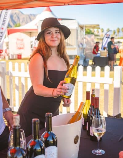 A woman in a black dress and wide-brimmed hat stands behind a table with various bottles of gourmet wine on it. She is smiling and holding a bottle of white wine, ready to pour it. There's a white picket fence and colorful tents in the background, suggesting an outdoor event in Palm Springs.