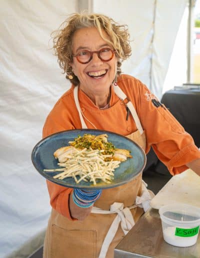 A smiling person with curly hair and round glasses, wearing an orange chef's jacket and an apron, stands in a Palm Springs tent holding a blue plate of gourmet food. The dish on the plate appears to involve sliced ingredients and a garnish. A container is on the table next to them.