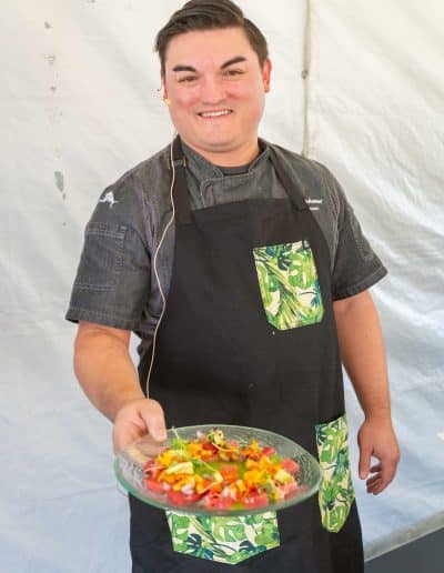 A person wearing a black apron with green leaf patterns smiles while presenting a gourmet dish on a glass plate. The background is plain white, evoking the elegance of a Palm Springs kitchen.