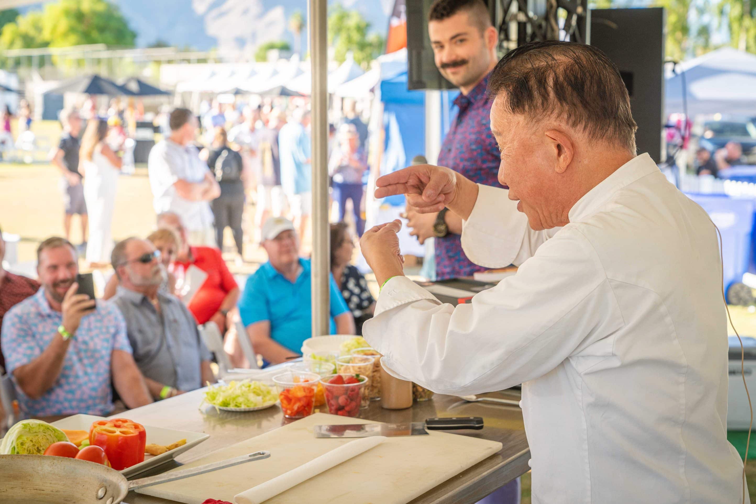 A chef, dressed in a white coat, demonstrates a cooking technique on stage at an outdoor Palm Springs event. Around him, people sit and watch attentively, some sipping wine. Various ingredients including vegetables are visible on the counter, set against a backdrop of mountains, tents, and blue sky.