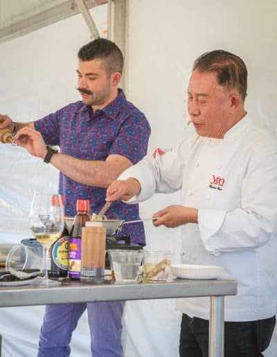 A chef in a pristine white coat and a man in a colorful shirt are performing a cooking demonstration on stage in Palm Springs. The gourmet chef holds a small bowl, while the other man pours a liquid. Various cooking ingredients and utensils are neatly arranged on the table.