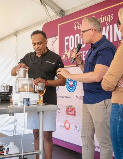 A chef uses a food processor on a table with kitchen equipment, while another man in glasses and a blue shirt holds a microphone to interview him. They are at the Palm Springs Food & Wine Festival, as indicated by the banner in the background.