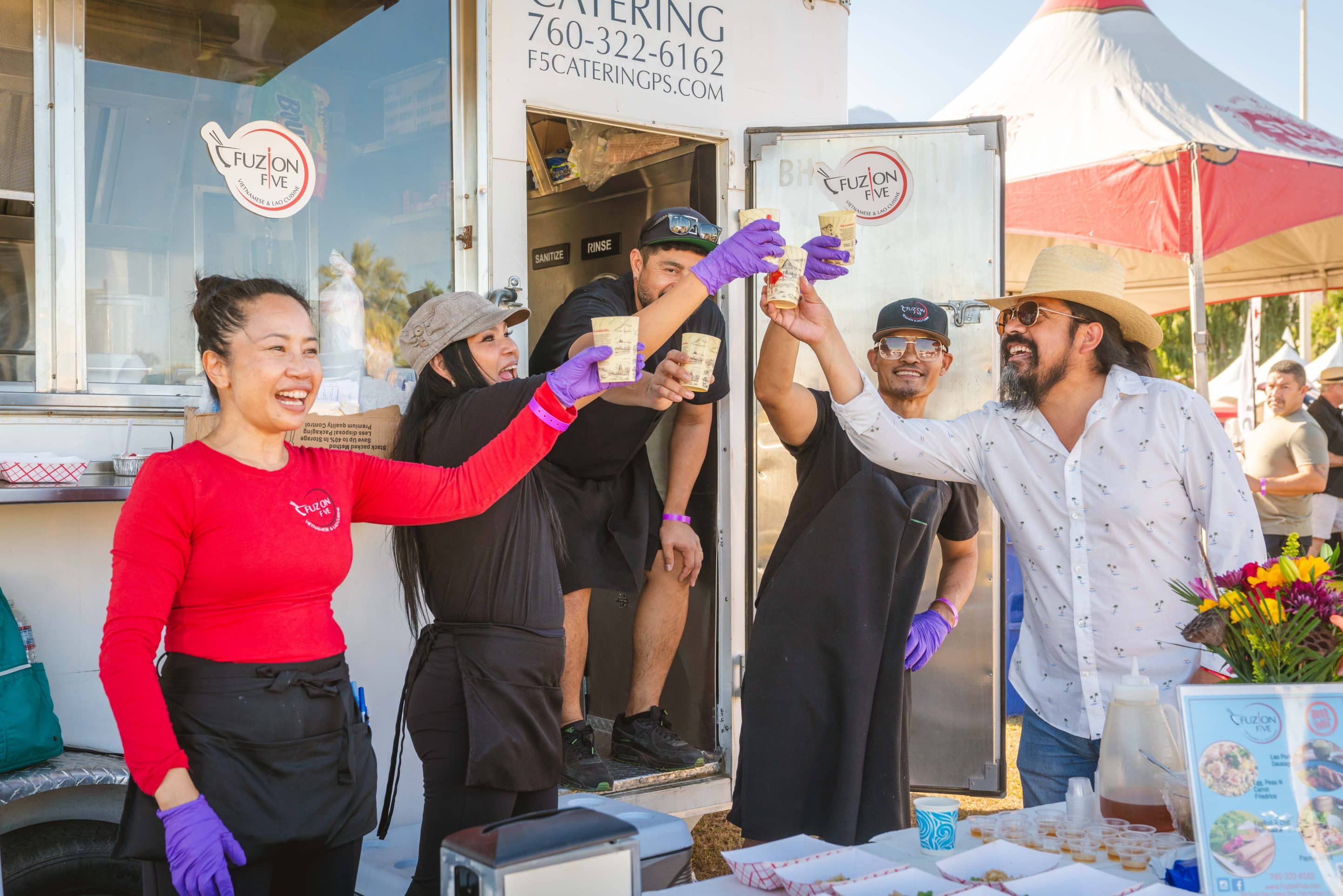 A group of people stands outside a food truck, raising cans in a celebratory toast. They are smiling and wearing casual clothing, including aprons and hats like seasoned chefs. One person holds a bouquet of yellow and orange flowers. A festival tent and other booths are visible in the background, adding to the gourmet atmosphere.