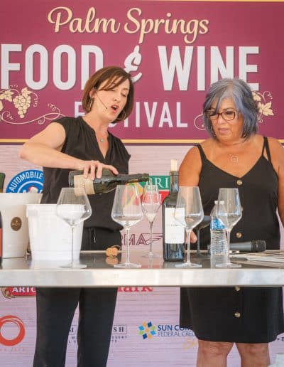 Two women are standing behind a table at the Palm Springs Food & Wine Festival. The woman on the left, like a gourmet chef, is gracefully pouring wine into glasses, while the woman on the right looks on appreciatively. The table is adorned with several wine glasses, water bottles, and wine bottles.