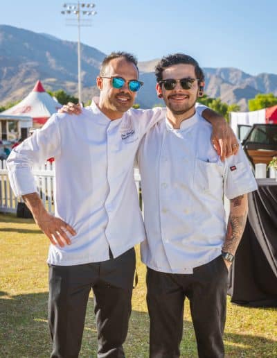 Two chefs in white uniforms and sunglasses stand smiling with mountains and tents visible in the background. One chef, holding a glass of wine, has his arm around the other. Both have short hair and are dressed in black pants, giving a gourmet touch to this outdoor event on a sunny day.