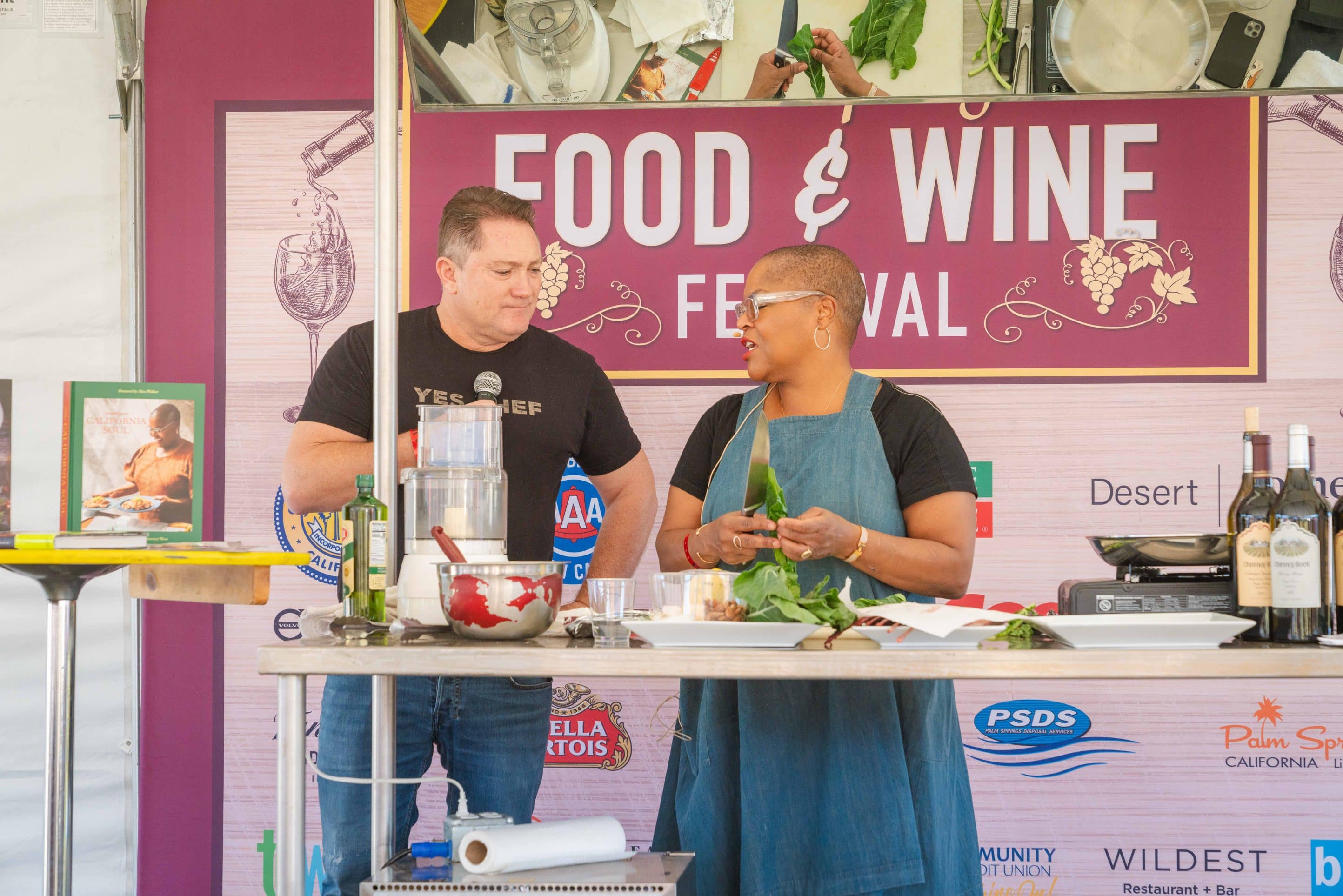 A man and a woman are cooking on stage at the Palm Springs Food & Wine Festival. The man, holding a microphone, speaks while the woman holds gourmet green vegetables. The stage is set with various kitchen utensils and bottles of wine, with a backdrop displaying "Food & Wine Festival.