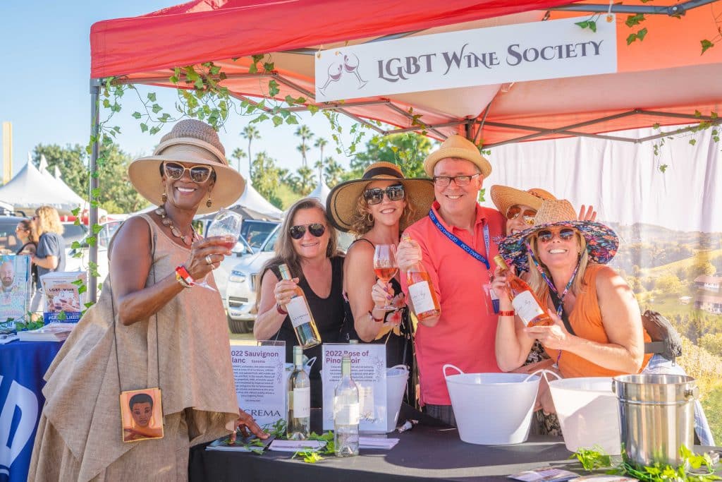 a vibrant culinary showcase in a bustling indoor venue, featuring elegantly arranged gourmet dishes and wine pairings, with enthusiastic attendees savoring and engaging with local chefs, all under warm, ambient lighting that captures the festive atmosphere of the palm springs food and wine festival. A group of people stand happily under a canopy labeled "LGBT Wine Society," holding bottles of gourmet wine. They are wearing summer hats and sunglasses, smiling at the camera. The booth is decorated with green vines, and various premium wine bottles are displayed on the table like a chef's curated selection.