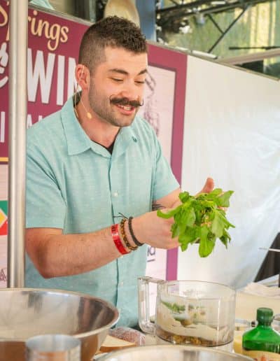 A bearded chef is preparing gourmet food at an outdoor cooking demonstration. He is holding fresh herbs and smiling, wearing a light blue short-sleeve shirt and a microphone headset. There are bowls, a food processor, various ingredients, and a bottle of wine on the table in front of him.