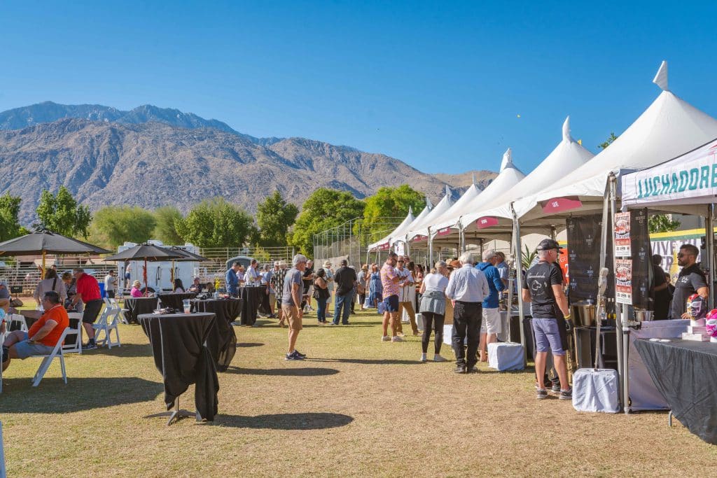 an elegantly set dining table adorned with gourmet dishes and fine glassware, framed by dramatic desert mountains under a vibrant sunset, epitomizing the luxurious yet relaxed atmosphere of a premier culinary event in palm springs. People enjoying an outdoor fair under white tents on a sunny day in Palm Springs. The fair is set on a grassy area with mountains in the background. Attendees are mingling, chatting, and visiting various booths featuring gourmet treats and wine. Tables and chairs are scattered around for seating.
