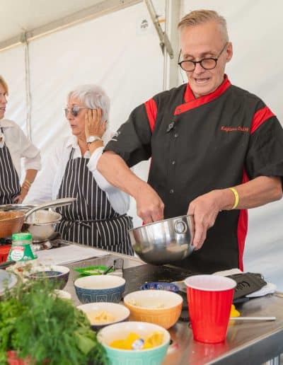 A man in a black and red chef's jacket is mixing ingredients in a bowl at a cooking demonstration in Palm Springs. He is focused on his task. Next to him, an older woman with white hair and glasses adjusts her glasses while wearing a black and white striped apron. Various colorful bowls are on the table.