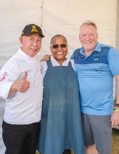 Three people are standing together and smiling for the camera in sunny Palm Springs. The person on the left is wearing a white chef's jacket and a black cap, giving a thumbs-up. The person in the middle is in an apron and sunglasses. The person on the right is in a blue polo shirt, holding a glass of wine. They are outdoors.