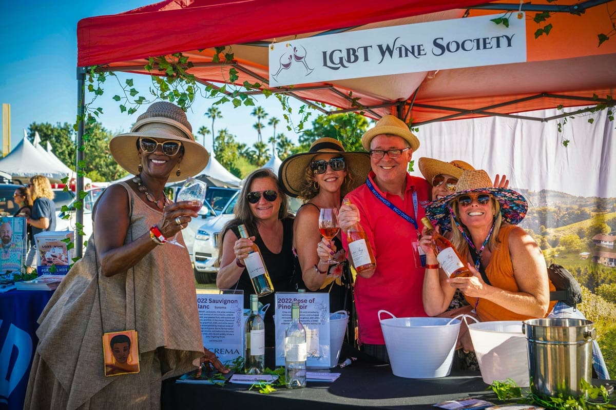 A group of five cheerful people, four women and one man, stand under a red tent with a sign that reads "LGBT Wine Society" at an outdoor event in Palm Springs. They are smiling, wearing summer outfits and hats, and holding bottles of wine. The background features more tents and people enjoying the gourmet atmosphere.