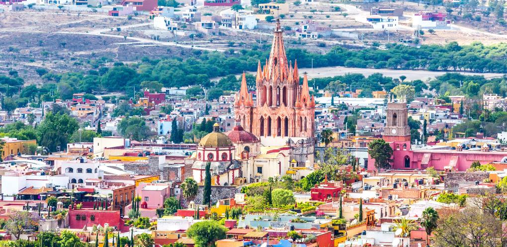 A scenic view of San Miguel de Allende in November showcases the colorful buildings and the iconic Parish of San Miguel Arcángel with its neo-Gothic architecture, surrounded by lush greenery and hills under a clear sky—a perfect backdrop for savoring local wine.